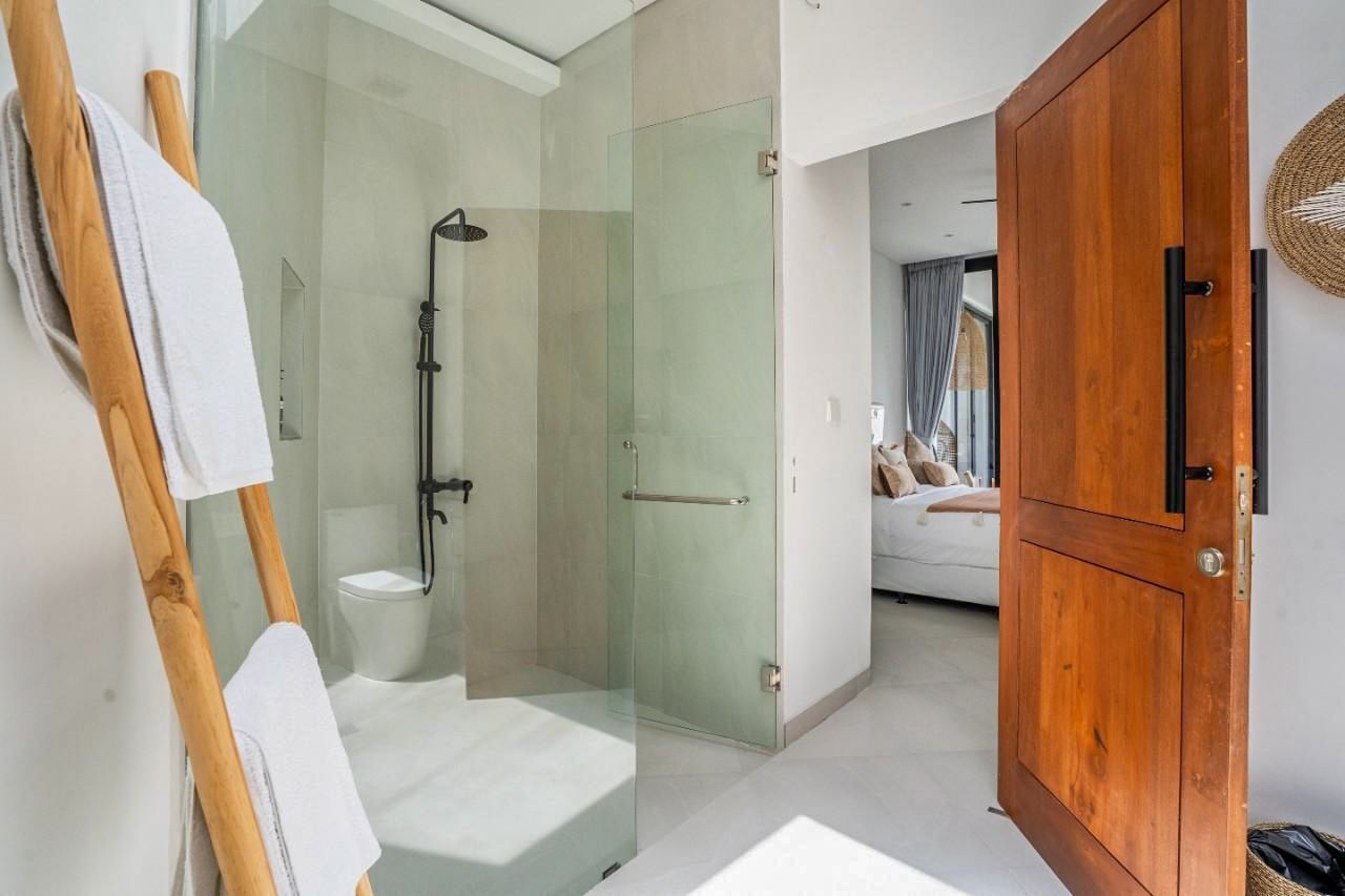 Um banheiro moderno com chuveiro em gabinete de vidro, um armário de madeira e uma cama visível no quarto adjacente.