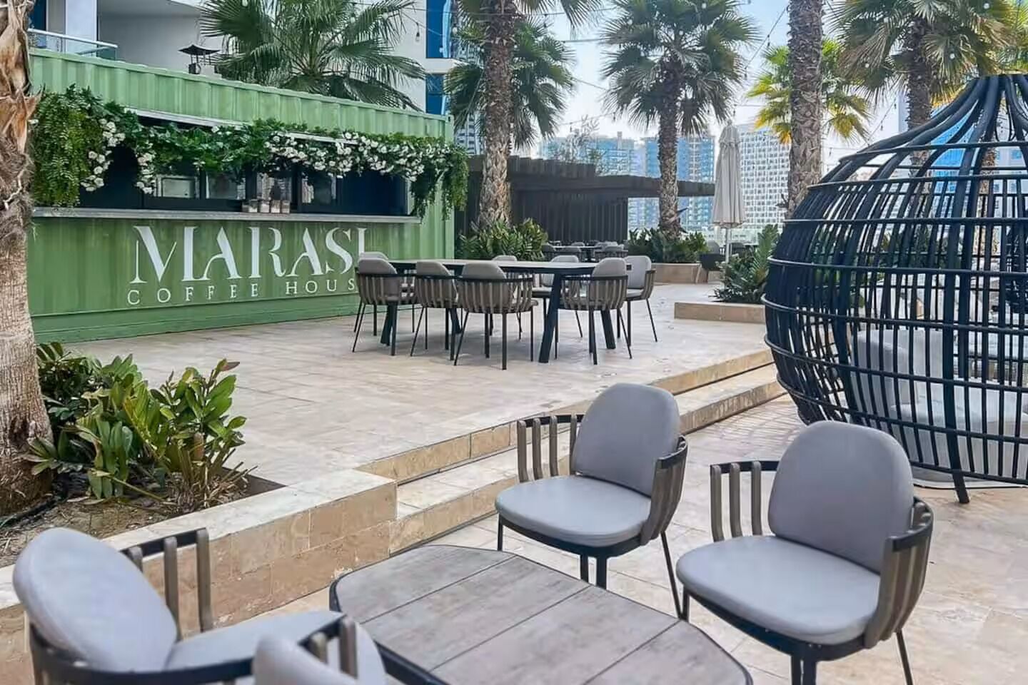 An outdoor seating area with tables and chairs, a green wall with 'MARAS COFFEE HOUSE' signage, and palm trees.