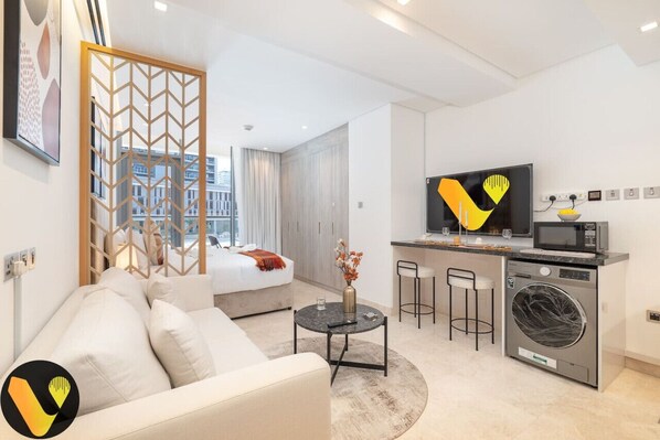 A modern hotel room with a bed, a sofa, a dining area with a TV, a washing machine, and a view of the city.