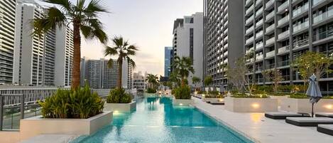 A rooftop swimming pool with lounge chairs, surrounded by high-rise buildings and palm trees.