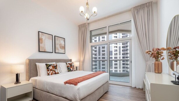 A modern bedroom with a large bed, a nightstand, a dresser, and a window with a city view.