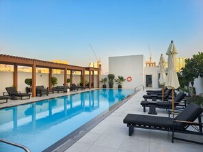 A rooftop swimming pool with lounge chairs and umbrellas, surrounded by potted plants and a clear sky.