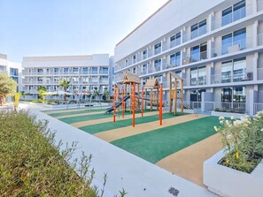 A multi-story building with balconies, a playground with slides and climbing structures, and a green play area.