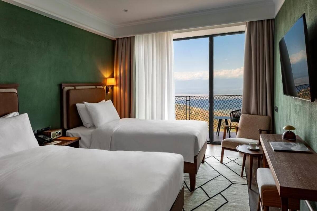 A hotel room with two beds, a desk, a chair, a TV, and a balcony with a view of the sea.