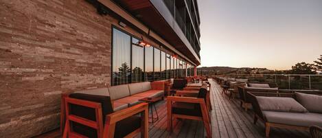 Una terraza de madera con asientos, ventanas amplias y vistas al paisaje.