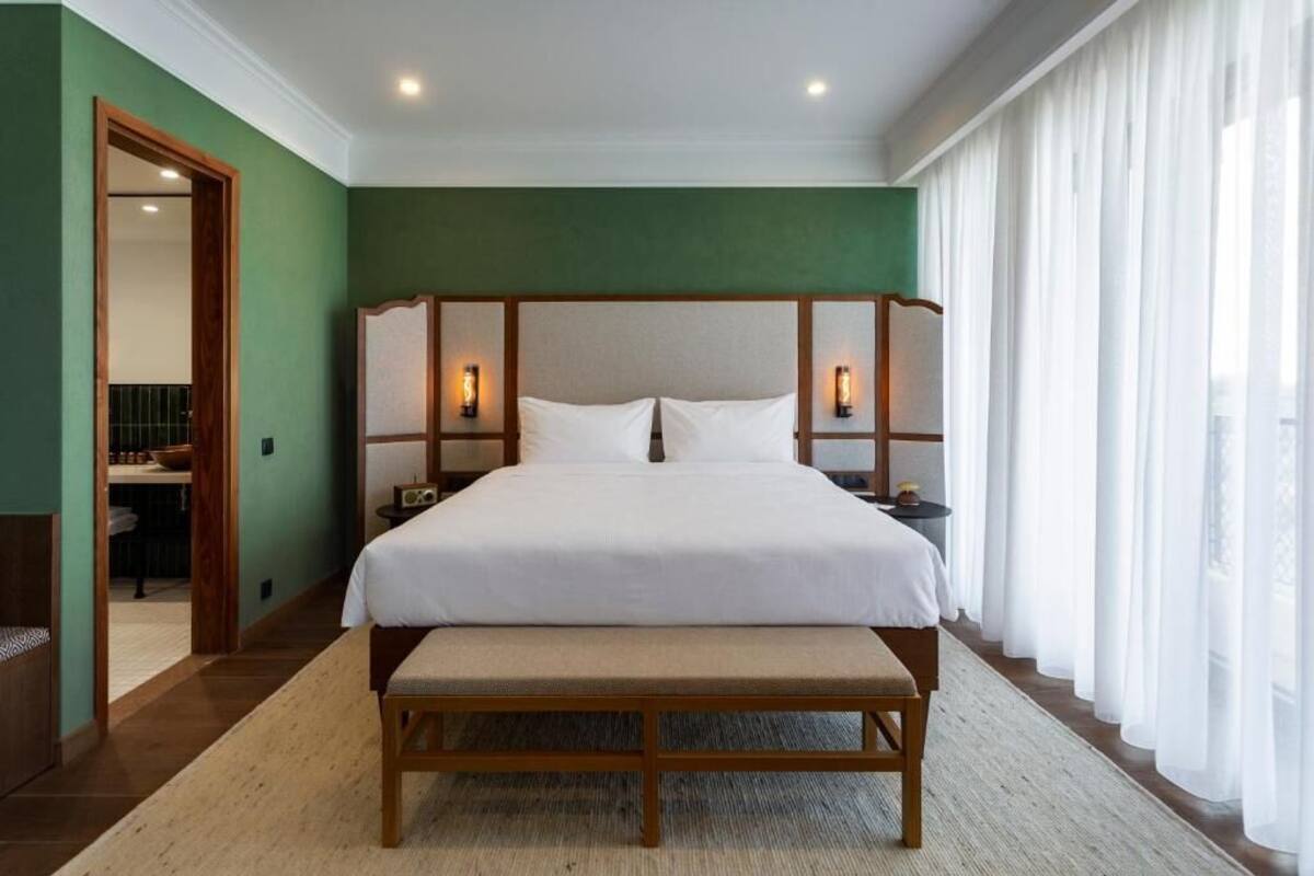 A hotel room with a large bed, wooden headboard, bedside lamps, a bench, and a view of the outdoors through sheer curtains.
