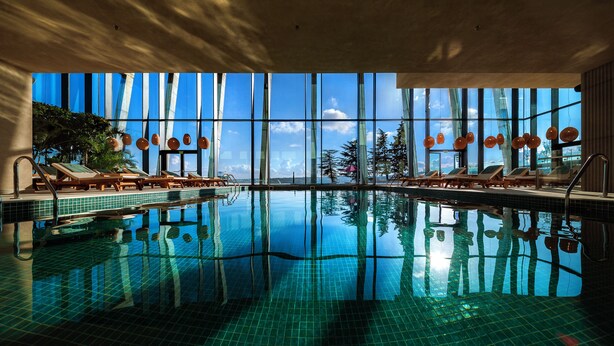 A large indoor swimming pool with clear reflections, sun loungers, and large windows offering a view of the outdoors. - BODBE Hotel (Sighnaghi)