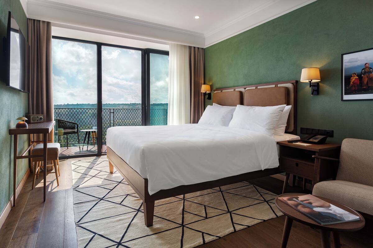 A hotel room with a large bed, a desk, a chair, a sofa, a TV, a lamp, and a balcony with a view.