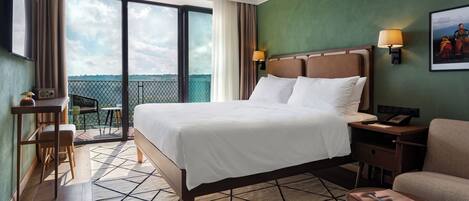 A hotel room with a large bed, a desk, a chair, a sofa, a TV, a lamp, and a balcony with a view.