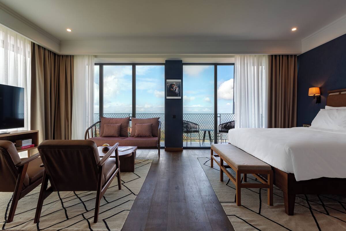 A hotel room with a bed, a sofa, a desk, and a TV. There is a large window with a view of the sky and clouds.