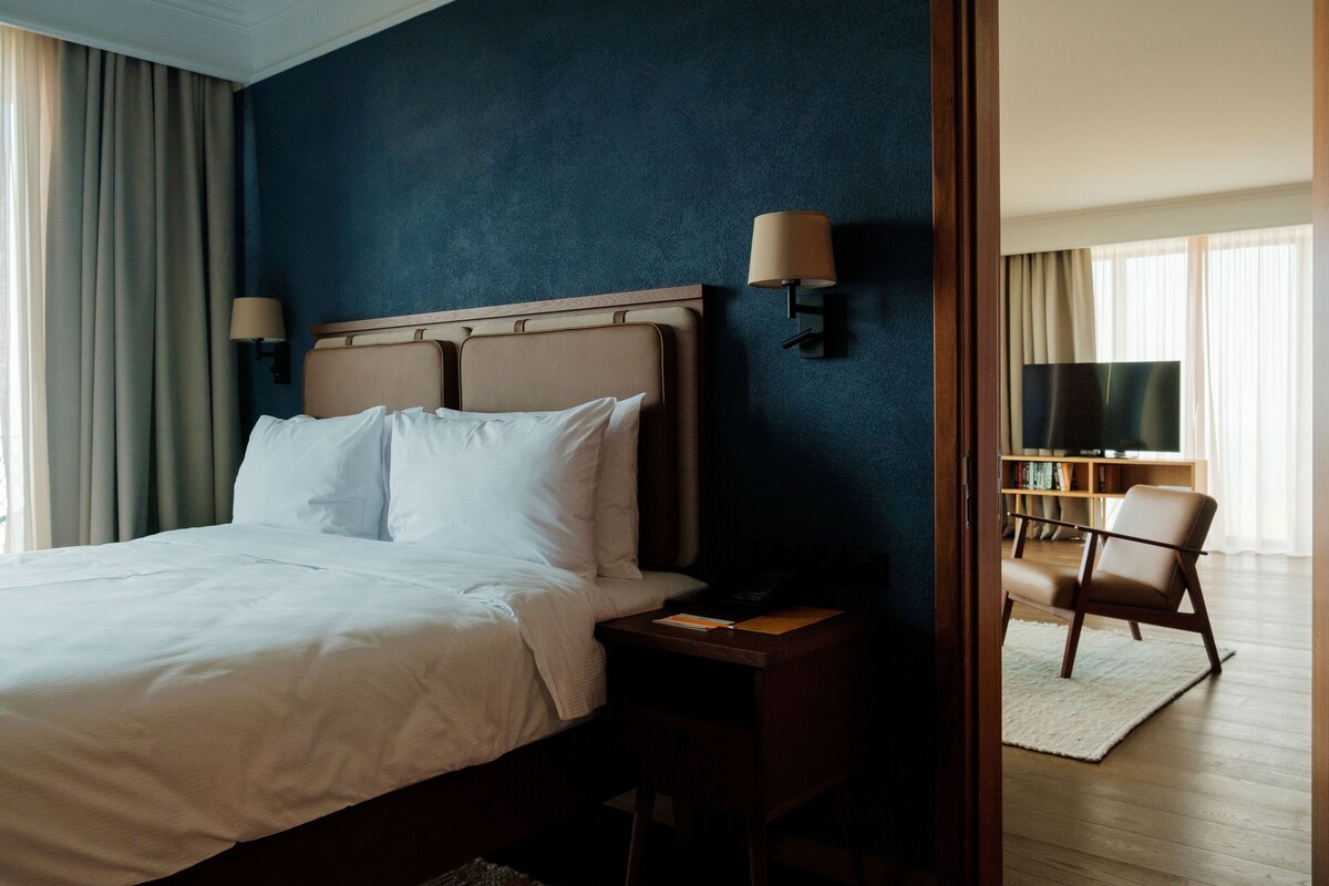 A hotel room with a large bed, two bedside lamps, a nightstand, a chair, and a television in the corner.