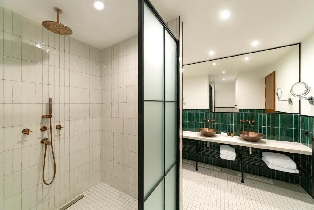 Une salle de bain moderne dotée d’une douche à parois de verre, d’un espace de douche carrelé de carreaux blancs et d’un meuble-lavabo recouvert de carreaux verts, avec deux vasques et un grand miroir.
