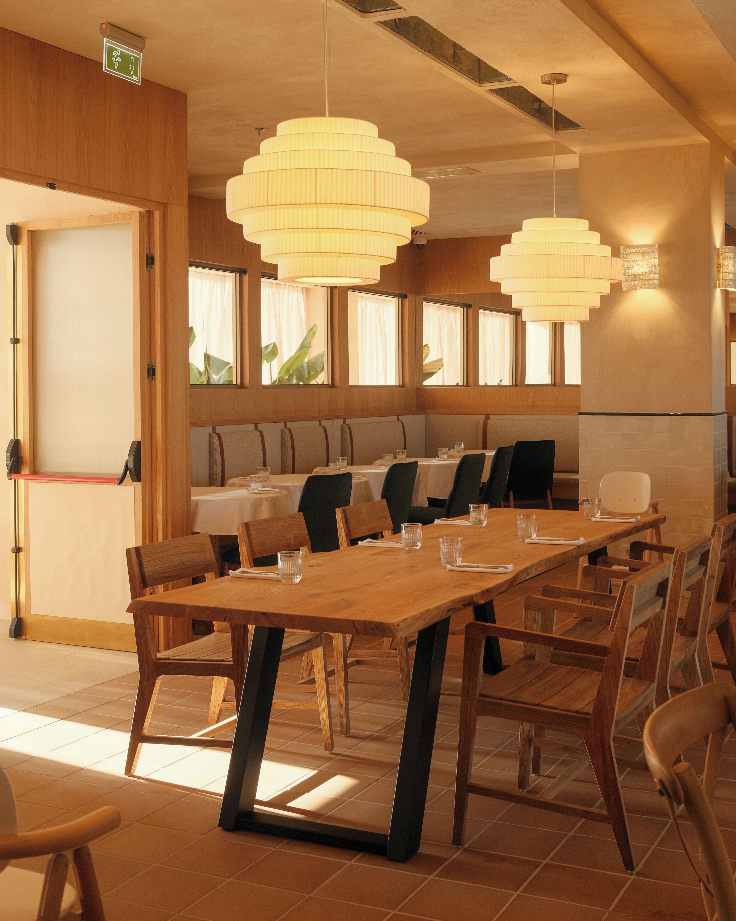 A restaurant with wooden tables and chairs, large windows, and hanging pendant lights.