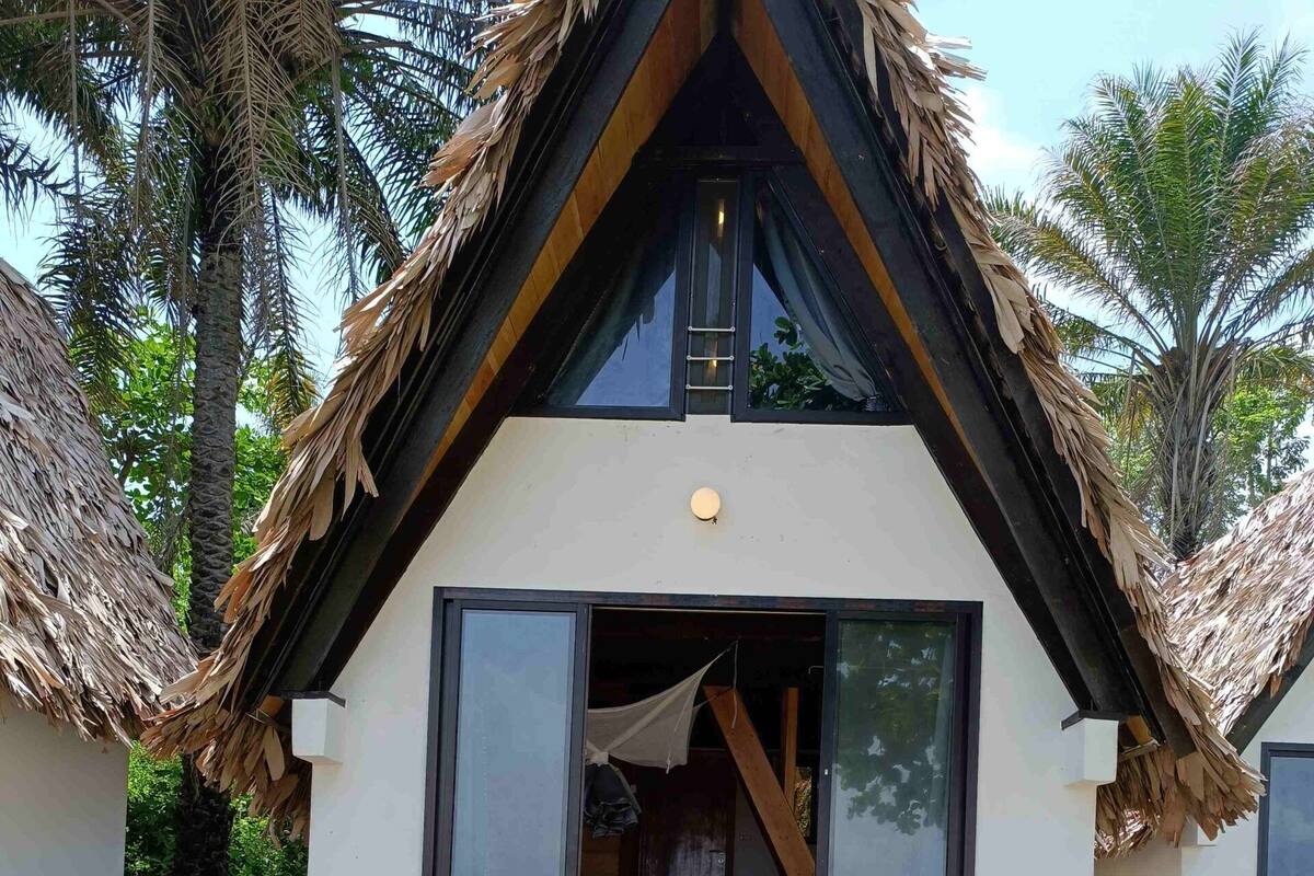 Un bungalow doté d’un toit de chaume, de grandes fenêtres et d’un escalier en pierre menant à un salon extérieur verdoyant.