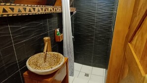 A small, wooden bathroom with a sink, a green trash bin, and a shower area with a black tiled wall.