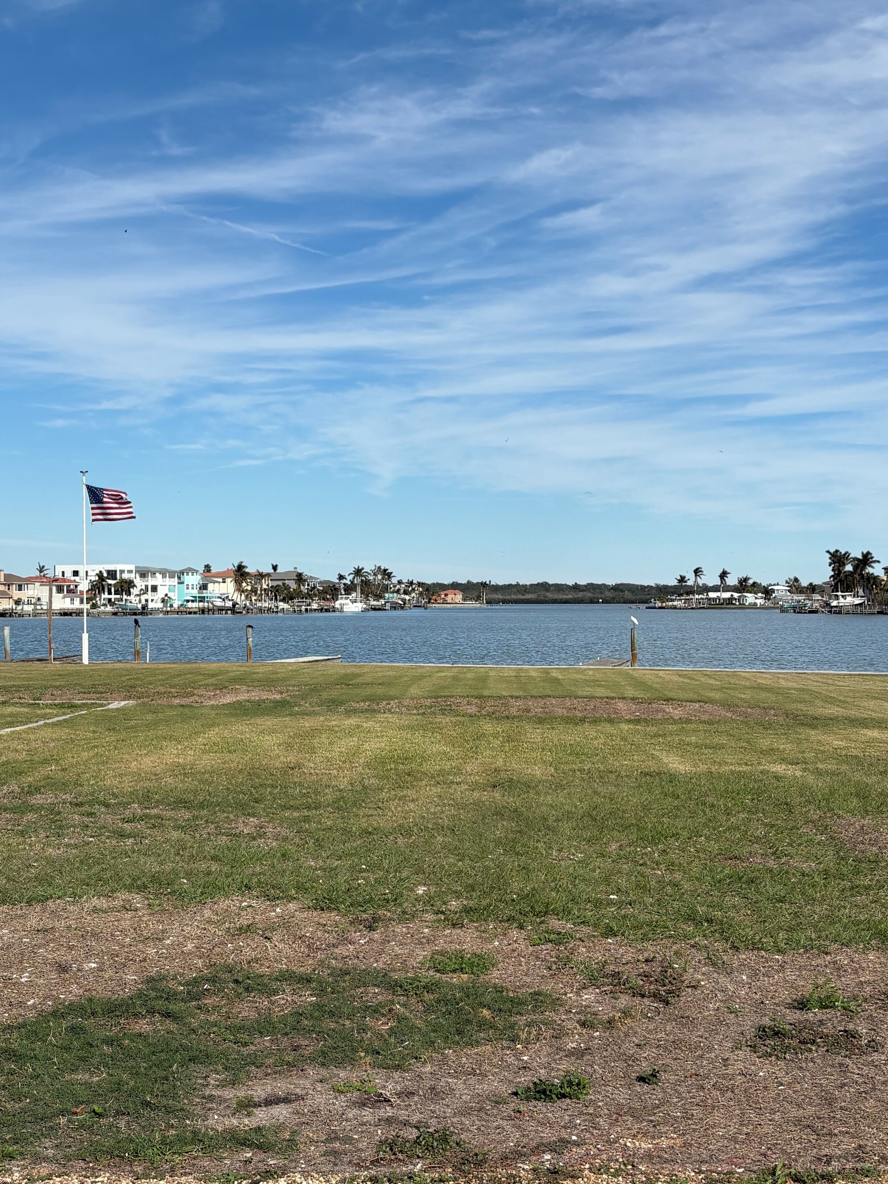 A grassy area with a flagpole and an American flag, a body of water, and a distant shoreline with buildings.