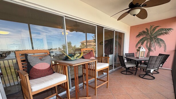 Un balcon avec des chaises et une table en bois, un ventilateur de plafond et une vue sur les palmiers et les immeubles.