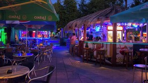 An outdoor bar area with tables and chairs, a thatched-roof bar, and neon signage.