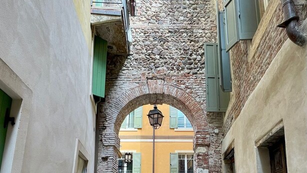 Un estrecho callejón con pavimento de adoquines, un arco de piedra y edificios con contraventanas verdes.