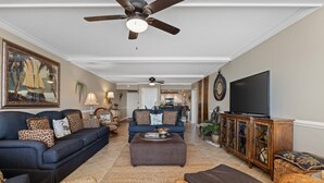 A spacious living room with a dark blue sofa, a brown ottoman, wicker furniture, a ceiling fan, and a flat-screen TV. - Upscale Oceanfront 3BR Condo • 14th Floor • Golf Course Views (Myrtle Beach)