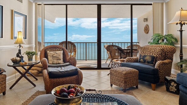 A spacious living room with a large sliding glass door leading to a balcony with a view of the ocean. There are wicker chairs, a sofa, and a coffee table with decorative items. - Upscale Oceanfront 3BR Condo • 14th Floor • Golf Course Views (Myrtle Beach)