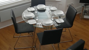 Una mesa de vidrio redonda con platos blancos, cubiertos y copas de vino, preparada para seis comensales.