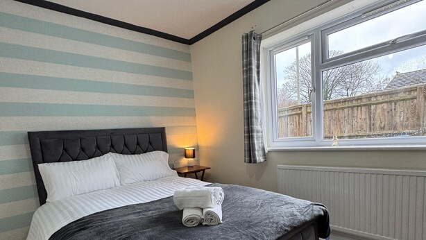 A neatly made bed with a dark headboard and a grey quilt, a bedside table with a lamp, and a window with a view of the outside. - Acaccia Bunglow  (Carterton)