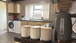 A kitchen with a washing machine, a kettle, and three jars labeled coffee, tea, and sugar. - Acaccia Bunglow  (Carterton)
