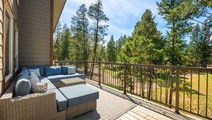 A covered outdoor seating area with a wicker sofa and cushions, a wooden deck, and a view of a forested area.
