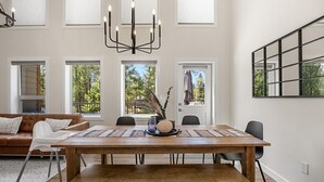A dining area with a wooden table, chairs, and a chandelier. There are large windows with blinds, a sofa, and a view of trees outside.