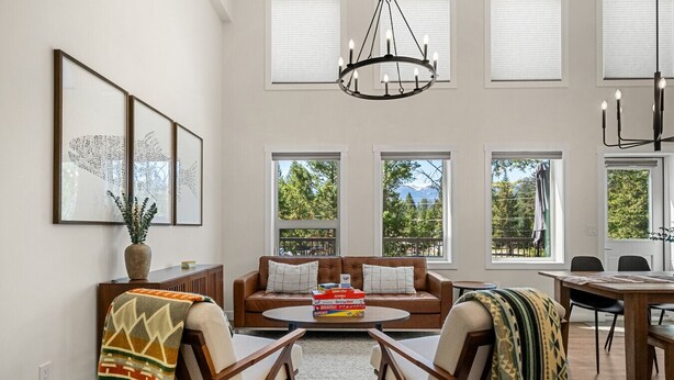 A spacious living room with a brown leather sofa, a coffee table, and a dining area with a wooden table and chairs. Large windows provide a view of the outdoors.