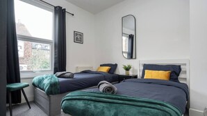 A hotel room with two beds, a window with curtains, a bedside table with a plant, and a mirror on the wall. - LFC Stay | 1 Min to Stadium | W/M | Pet Friendly (Anfield)