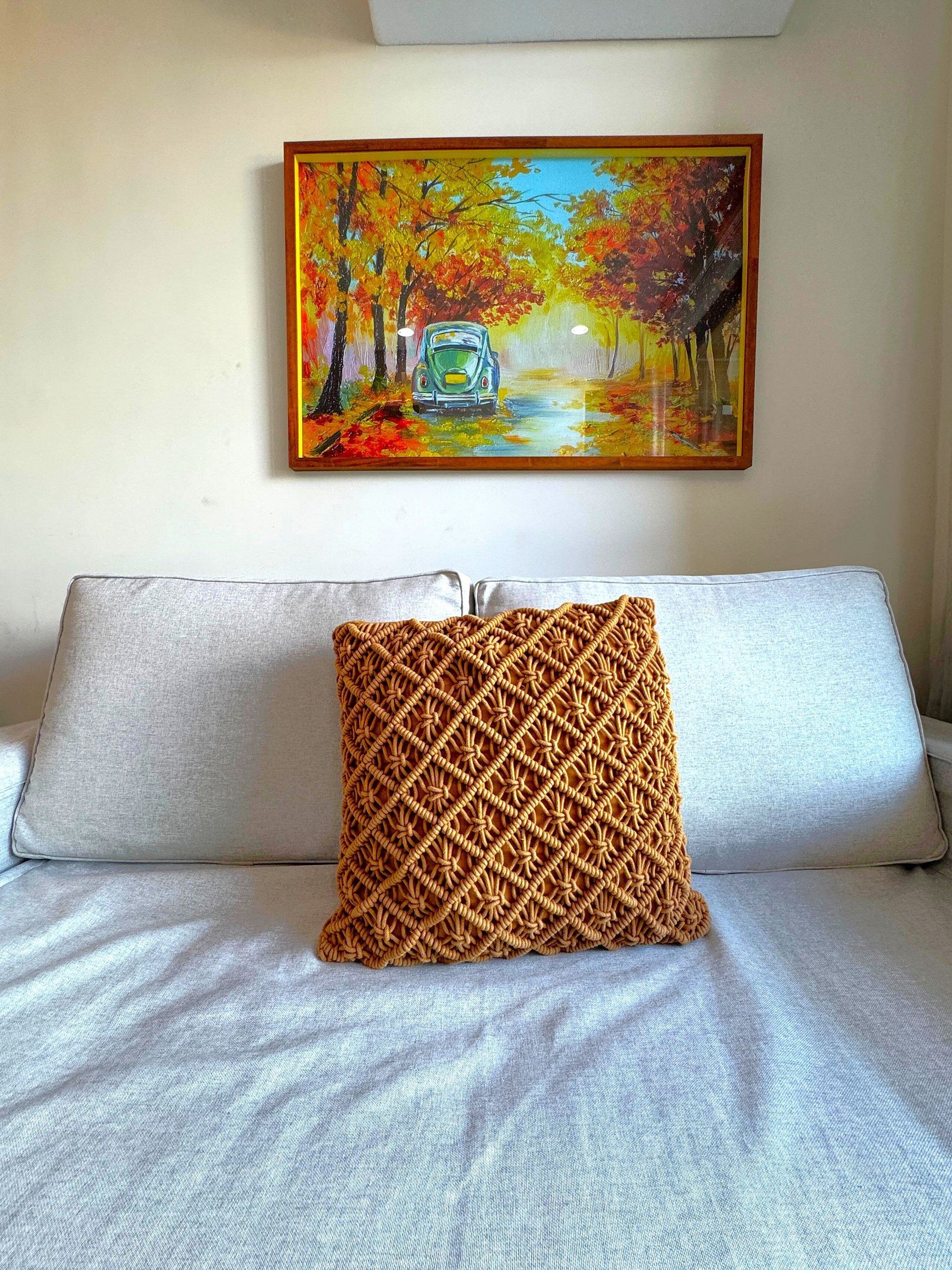 A bed with a patterned pillow and a painting of a car on a tree-lined road above it.