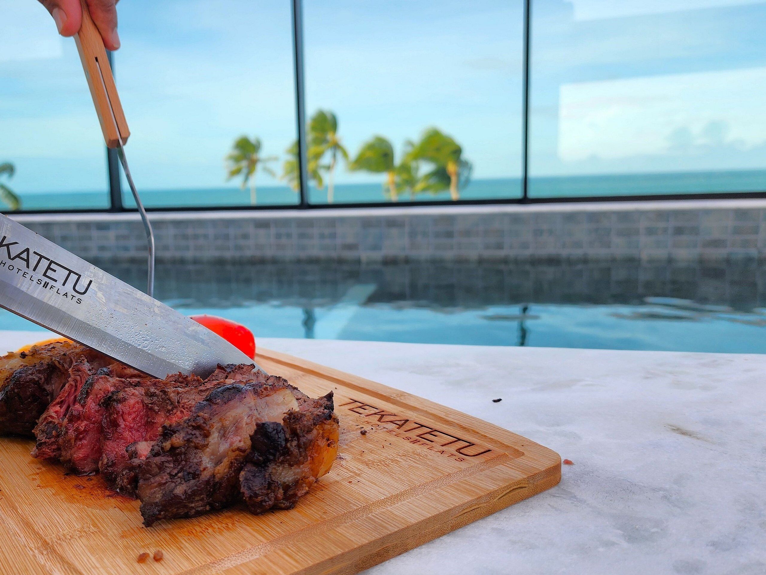 A steak being sliced on a wooden cutting board with a knife labeled "KATETU HOTELS FLATS" in a poolside setting with a view of palm trees and the ocean.