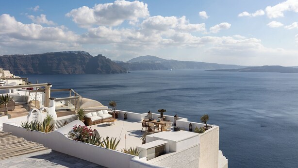 A rooftop terrace with a view of the sea, white buildings, and a distant mountain range.