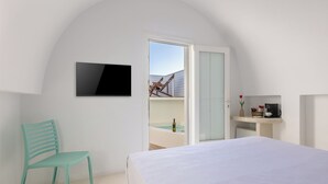 A bedroom with a bed, a TV mounted on the wall, a chair, a small table with a coffee machine, and a view of the outside balcony. - Santorini Sun Villa Two in Oia | Private Hot Tub (Oia)