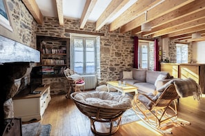 Een gezellige kamer met een stenen muur, houten balken, een boekenplank, een bank, een schommelstoel en een open haard.