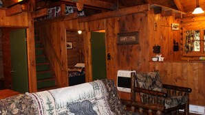 A cabin interior with a wooden staircase, a couch with a bear design, a chair, and a small kitchen area. - Cozy Log Cabin in Cook Forest ~ Dog Friendly (Clarington)