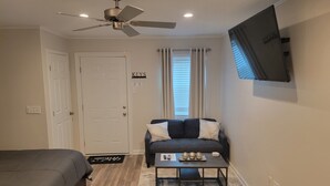 A compact hotel room with a bed, a small sofa, a coffee table, a ceiling fan, and a TV mounted on the wall. - Beautiful Studio Apt 10 - Upstairs (Baton Rouge)