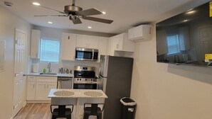 A compact kitchen with white cabinets, a microwave, a stove, and a refrigerator. There is a ceiling fan, a window with blinds, and a small trash bin. - Beautiful Studio Apt 10 - Upstairs (Baton Rouge)