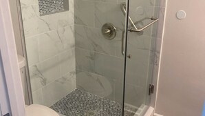 A glass-enclosed shower with a marble-tiled wall, a white toilet, and a wooden floor. - Beautiful Studio Apt 10 - Upstairs (Baton Rouge)
