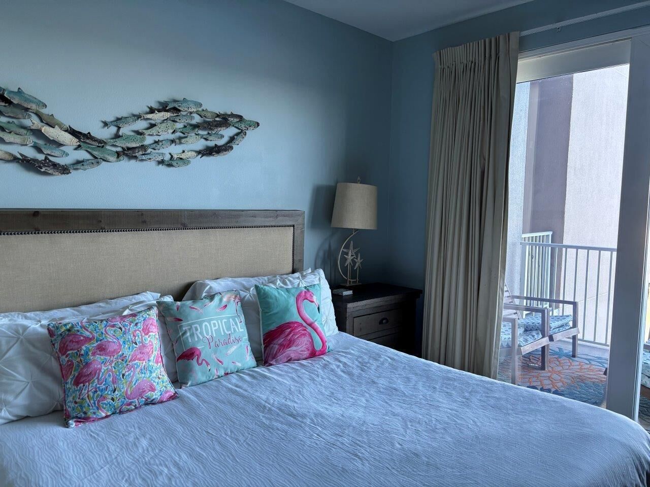 Een hotelkamer met een bed, kussens, een nachtkastje, een lamp en uitzicht op een balkon.