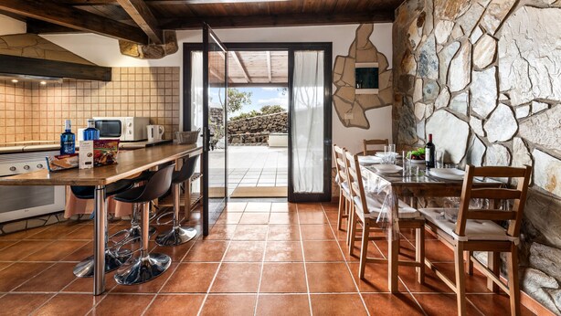 Une cuisine avec un mur en pierre, un coin repas avec des chaises, une table dressée pour deux personnes et une vue à travers une porte vitrée.