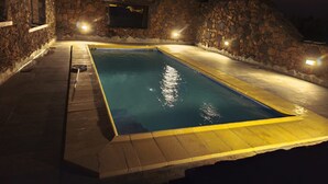 Une piscine rectangulaire entourée d’un mur en pierre, illuminée par des lampes fixées au mur.