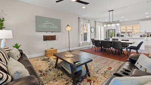 A modern living room with a sofa, coffee table, and a dining area with chairs and a table. There is a kitchen in the background with white cabinets and a window. - 3 Stry Treehouse Chickamauga 20 min to Chatt Vistas (Walker County)