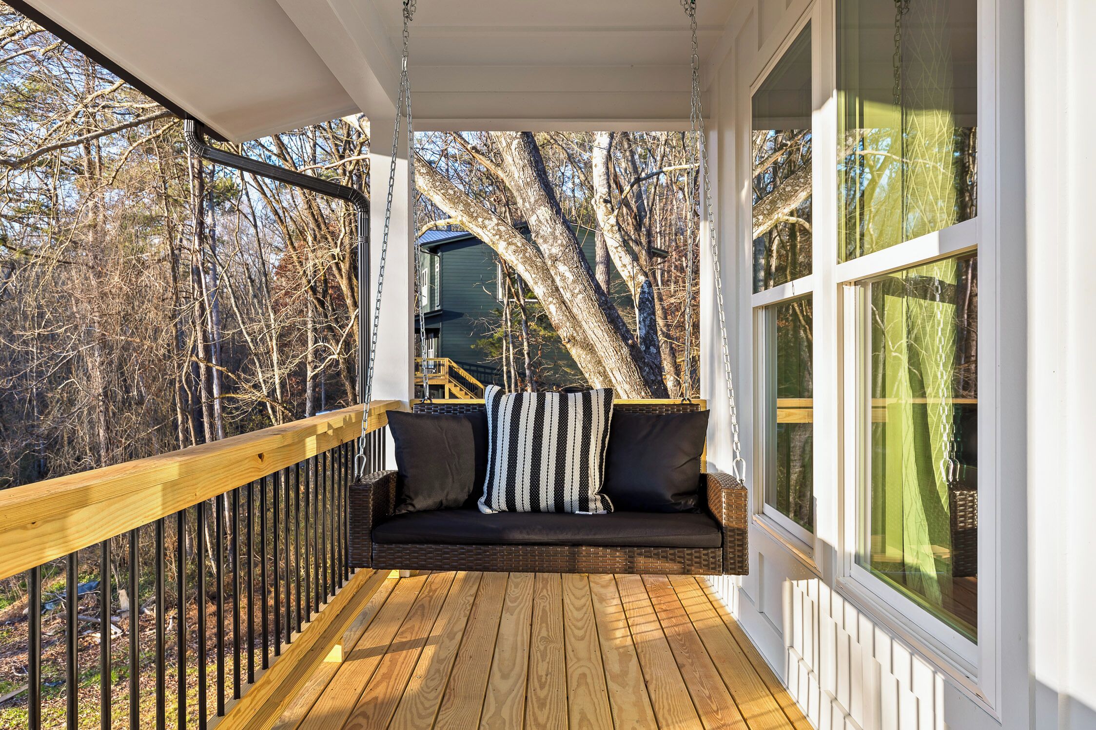 A wooden deck with a cushioned seating area, a railing, and large windows offering a view of trees and a house.