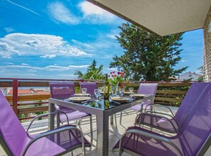 Um terraço com uma mesa posta para dois, rodeado por cadeiras roxas, com vista para uma paisagem com árvores e céu claro.