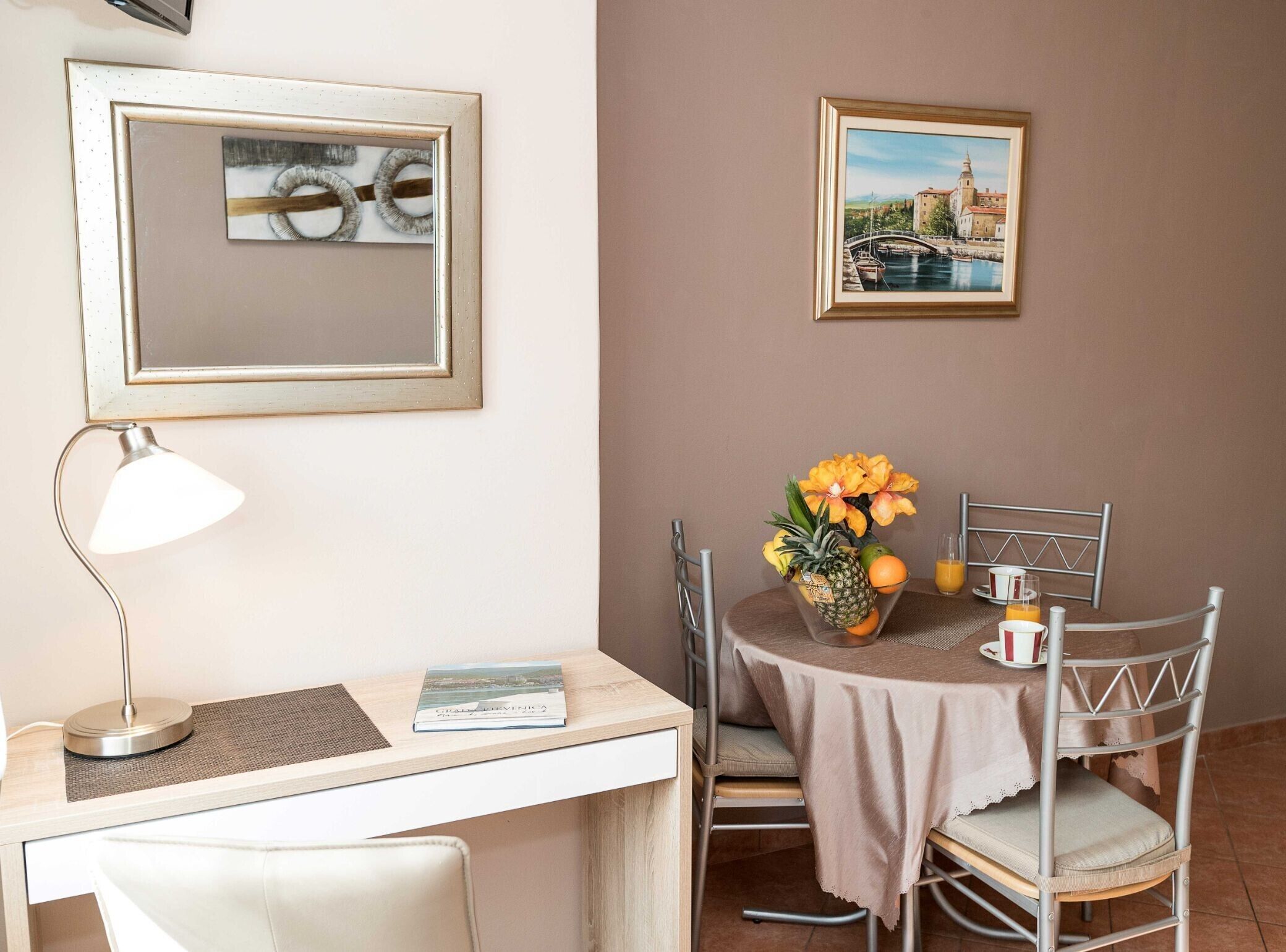 Een eethoek met een gedekte tafel voor twee personen, een lamp, een lijst met een afbeelding en een boek op tafel.