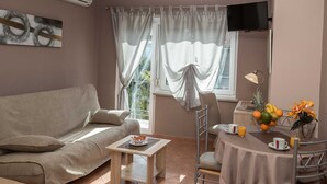 A modern, well-lit room with a sofa, a dining table with chairs, a small table with a bowl of fruit, and a TV mounted on the wall. - Beachfront Crikvenica Apartments with Sea View - Standard Studio Apartment with Sea View (Crikvenica)