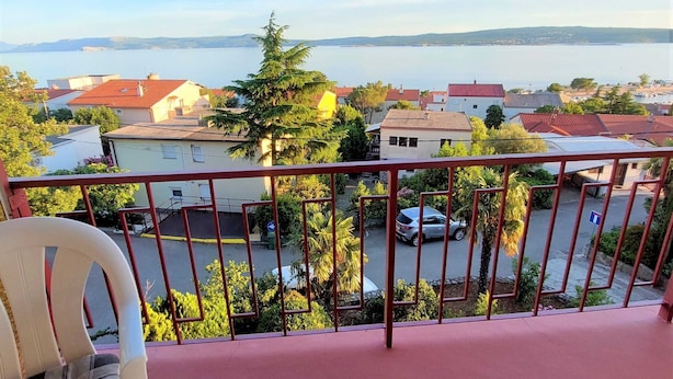 Un balcon avec une balustrade rouge donnant sur un quartier résidentiel, avec des maisons, une voiture et une étendue d’eau au loin.